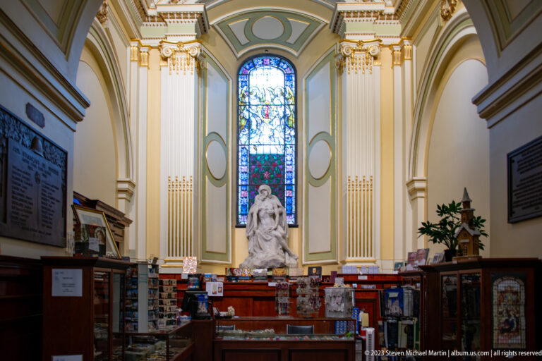 White religious statue beneath a tall stained-glass window, framed by cream-and-gold pilasters above cabinets and devotional displays.
