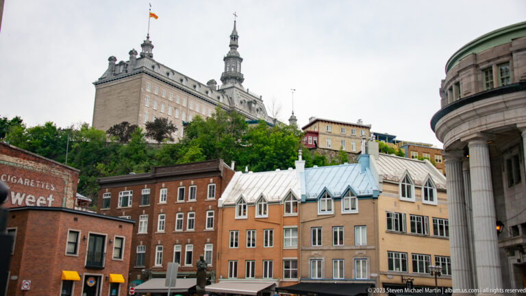 Historic Quebec City buildings climb a steep slope toward a tall tower, with a curved columned stone façade at right.