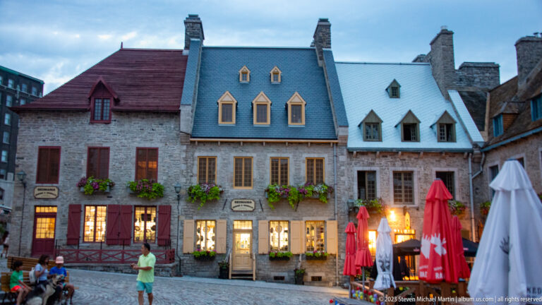 Three adjoining historic stone houses with red and blue-gray roofs, dormers, chimneys, shutters, flower boxes, and café umbrellas.