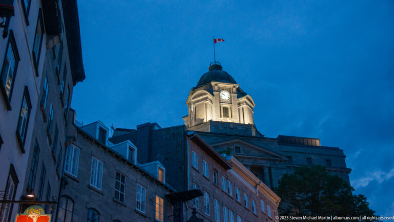 Illuminated domed clock tower with Canadian flag rising above dark historic façades against a deep blue evening sky.