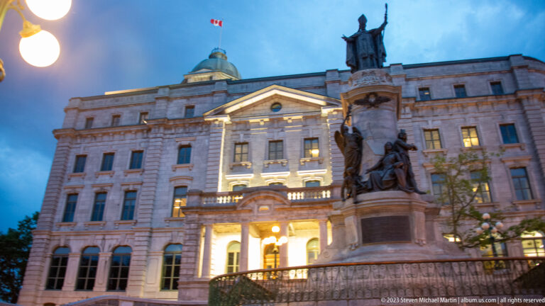 Large sculptural monument before an illuminated classical civic building with pediment, columns, dome, and Canadian flag at dusk.