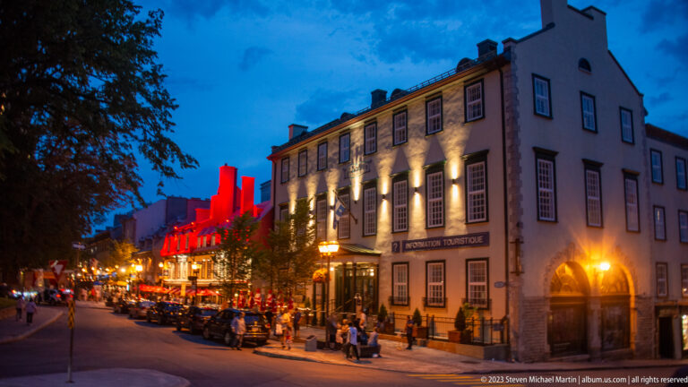 Hôtel Clarendon on a brightly lit corner with restaurants, pedestrians, outdoor dining, and red-lit historic façades extending down the street.