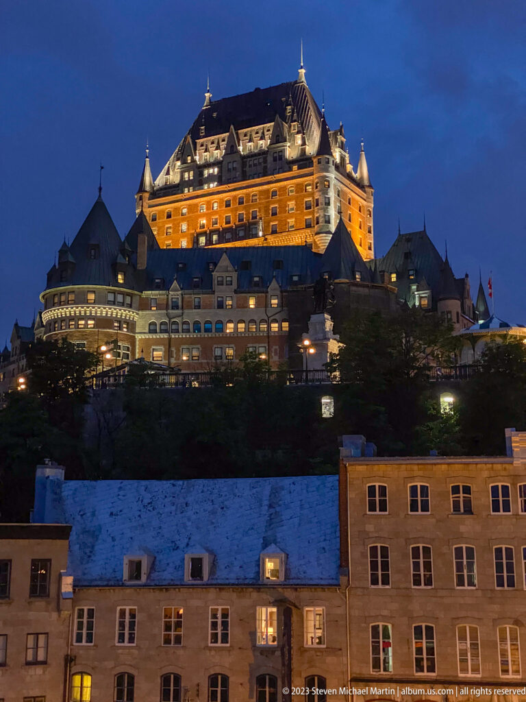 Illuminated Château Frontenac glowing orange on the ridge above dark terraces and Lower Town buildings under a deep blue sky.