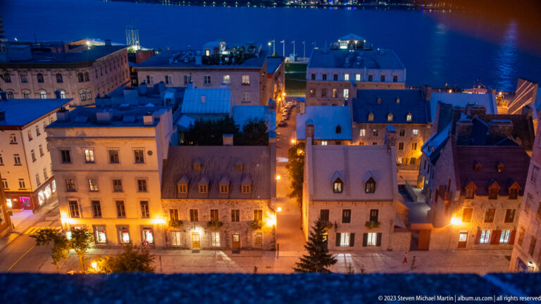 Elevated night view over illuminated Lower Town buildings and narrow streets toward the dark St. Lawrence River.