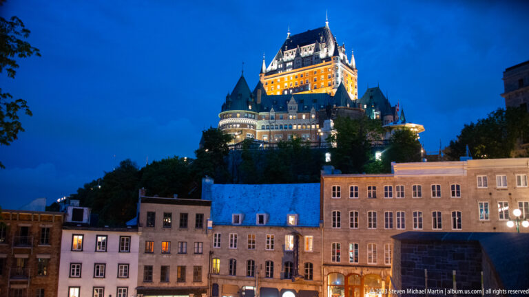 Château Frontenac illuminated above rows of Lower Town buildings, glowing windows, trees, and streetlights at night.