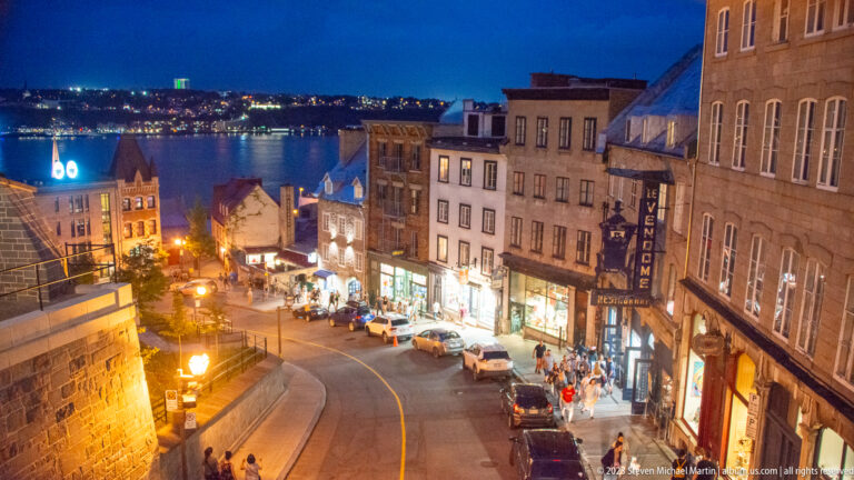 Curving illuminated Lower Town street descending past shops and parked cars toward the St. Lawrence River and distant shoreline lights.
