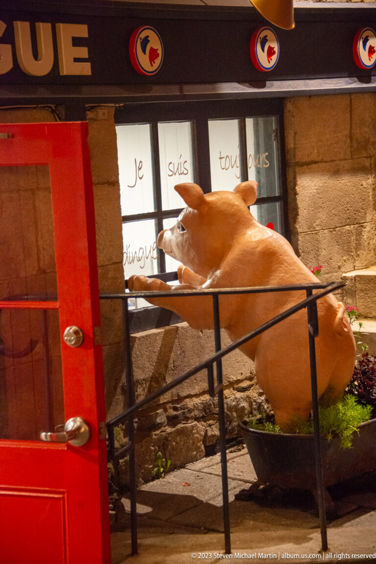 Large tan pig sculpture behind a black railing beside a bright red door and window with handwritten French words.