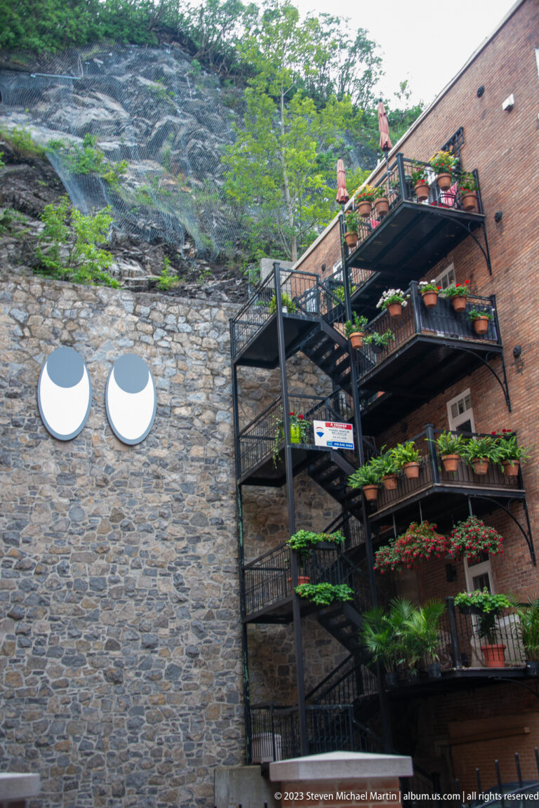 Black metal staircase zigzags upward beside a rough stone wall and brick building, with planters and people above.