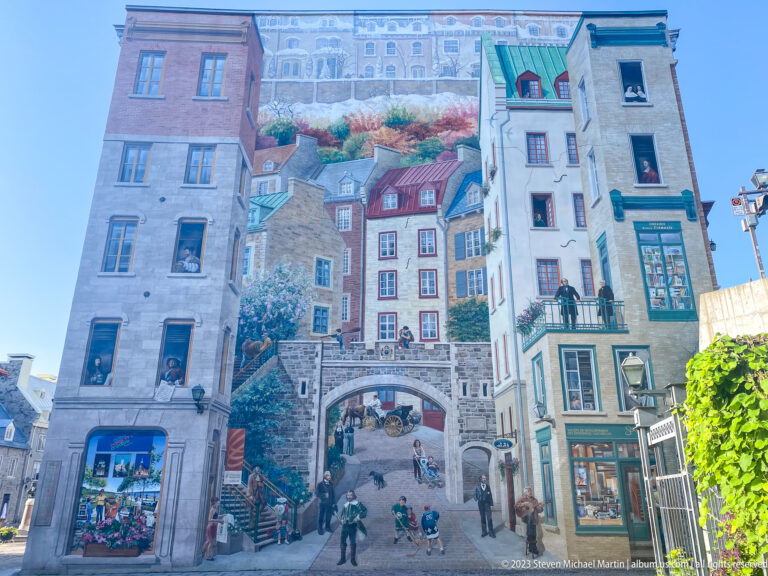 Large Fresque des Québécois mural covering a tall façade with painted houses, stairs, figures, gardens, windows, and central arch.