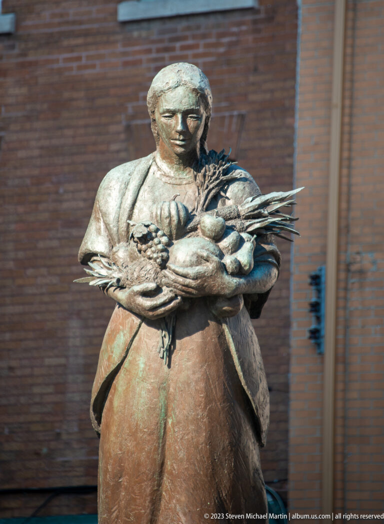 Bronze statue of a woman holding a large basket filled with vegetables, fruit, and leafy produce against her body.
