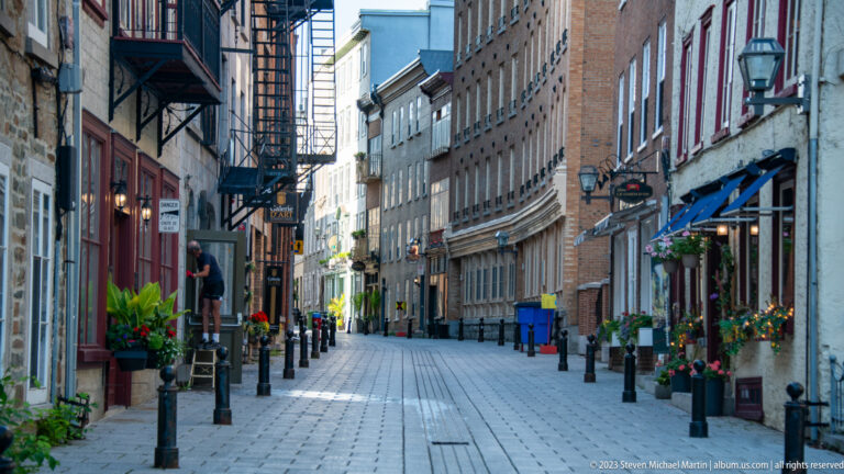 Narrow paved pedestrian lane lined with brick and stone shops, hanging signs, flowers, awnings, bollards, and a few people.