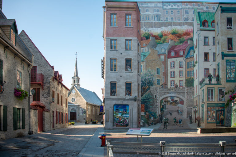 Fresque des Québécois mural beside historic stone buildings, with Notre-Dame-des-Victoires and its spire visible across Place Royale.