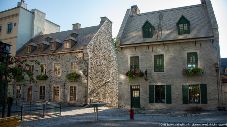 Adjoining historic stone houses with steep roofs, dormers, chimneys, green shutters, flower boxes, and varied pale masonry.