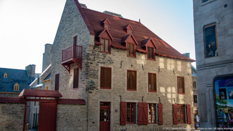 Historic stone house with steep red roof, three dormers, red shutters, small balcony, chimneys, and ground-floor doors.