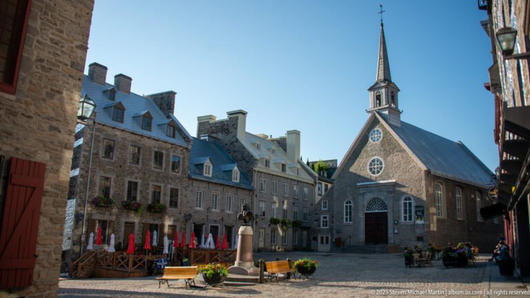 Notre-Dame-des-Victoires centered in Place Royale, with stone façade, round windows, tall spire, café umbrellas, planters, and surrounding historic buildings.