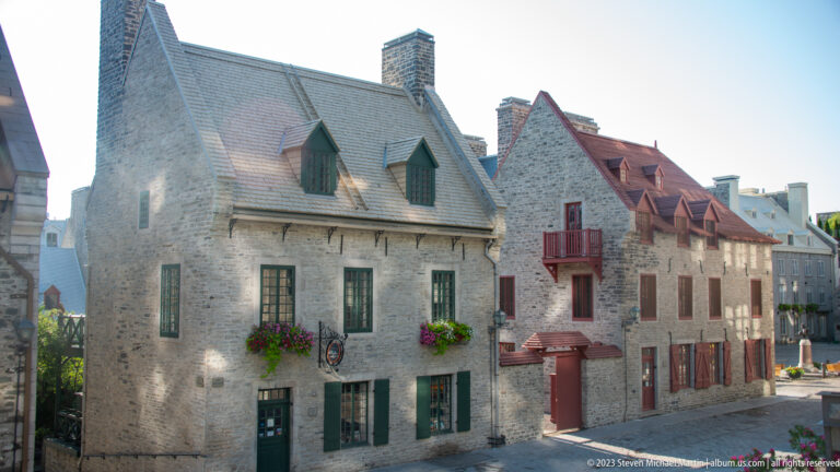 Two adjoining stone houses with contrasting gray and red roofs, green and red trim, dormers, chimneys, flower boxes, and balcony.