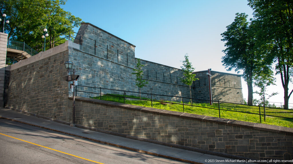 Massive gray stone fortification wall rising beside a sloping street, with stepped masonry, narrow openings, grass, railing, and trees.