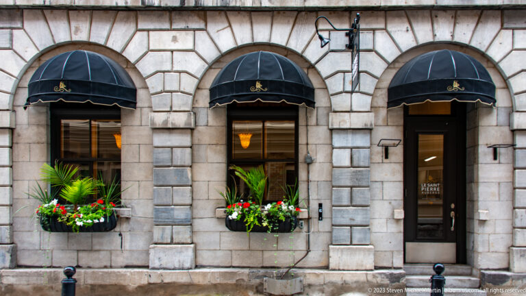 Three arched openings in a gray stone façade with black awnings and flower boxes filled with palms and red-and-white blooms.
