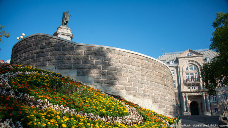 Curved gray stone rampart behind a sloping multicolored flower bed, with part of an ornate building visible beyond the wall.