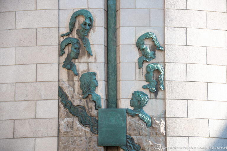 Several green-patinated bronze profile heads arranged vertically on a pale stone wall around a small rectangular plaque.