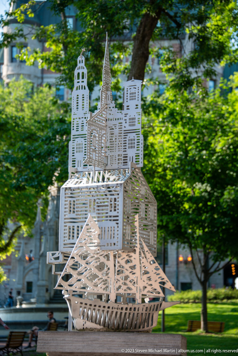 White openwork sculpture forming a church, tower, roofs, and surrounding buildings on a pedestal among green trees.
