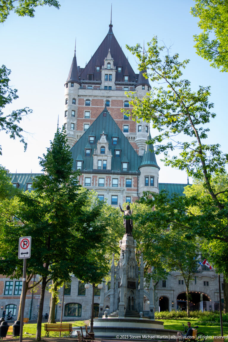Château Frontenac rising behind trees and a stone monument topped by a bronze figure in a landscaped public space.