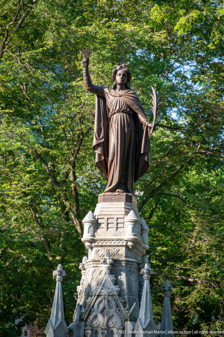 Robed bronze woman atop a carved stone monument, raising one hand and holding a palm frond against green trees.