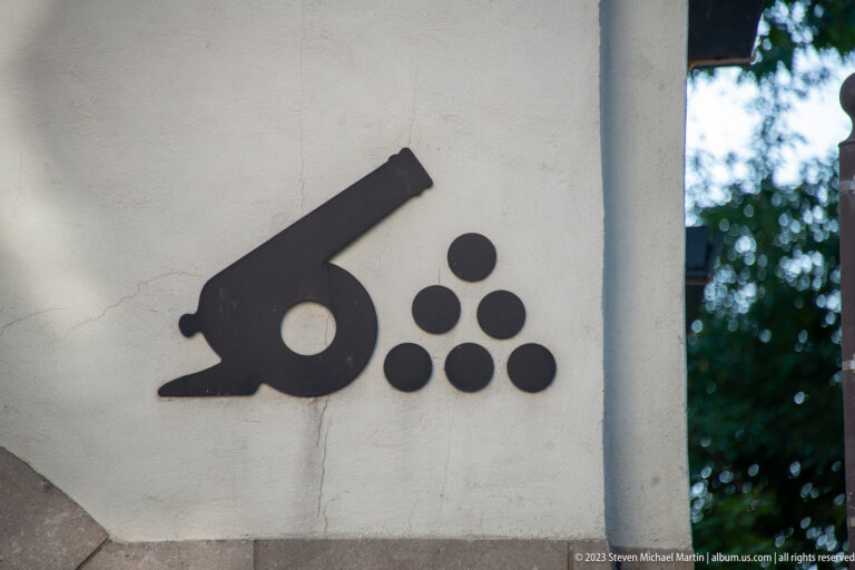 Black silhouette of a cannon beside six round cannonballs mounted on a pale exterior wall.