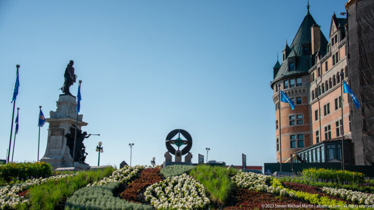 Formal flower beds with a tall monument, circular geometric sculpture, flags, and Château Frontenac under a clear blue sky.
