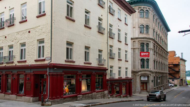 Pale corner building with dark red storefronts opposite an ornate rounded stone building on a narrow Quebec City street.