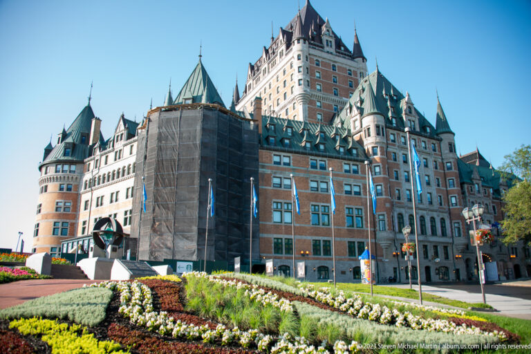 Château Frontenac above patterned flower beds, with copper roofs, turrets, tall central tower, and construction mesh on part of the façade.