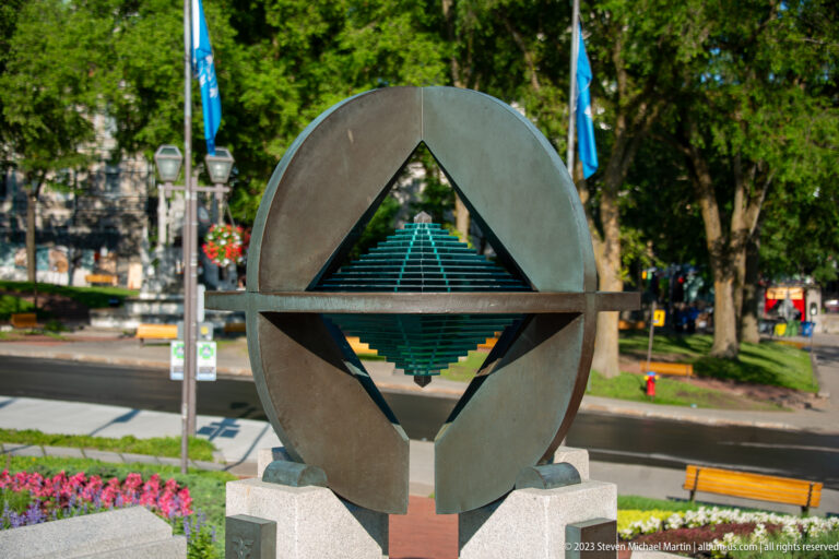 Dark circular metal sculpture containing a suspended green diamond-like geometric form, mounted on a stone base beside gardens.