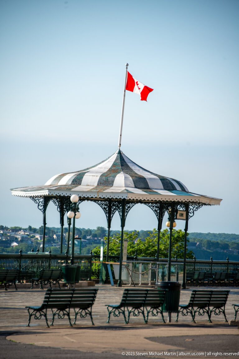 Ornate striped-roof gazebo with Canadian flag and empty benches on Dufferin Terrace overlooking the distant river landscape.