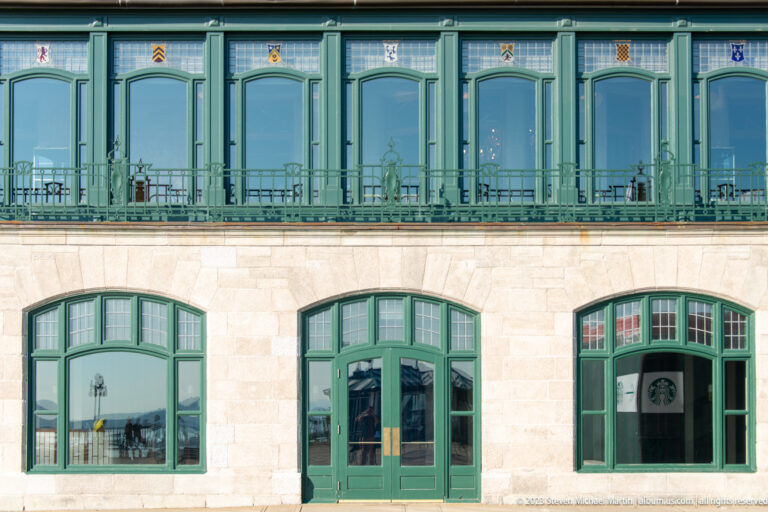 Pale stone building with green-framed arched windows and doors below a continuous glazed green-trimmed upper gallery.