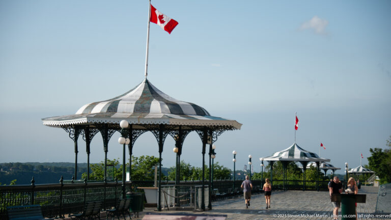 Multiple striped gazebos topped with Canadian flags recede along Dufferin Terrace as visitors walk beside the overlook railing.