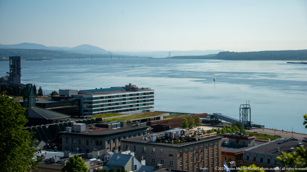 Wide elevated view over waterfront buildings to the St. Lawrence River, distant bridge, wooded shoreline, and hazy hills.