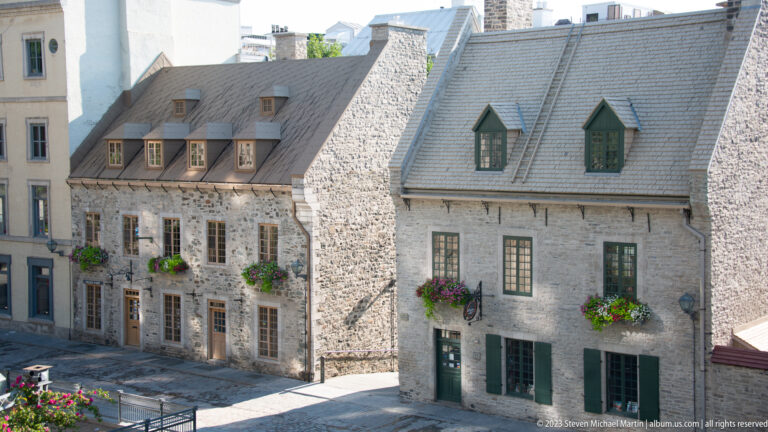 Two adjoining historic stone houses with steep gray roofs, chimneys, dormers, green windows, and flower boxes.