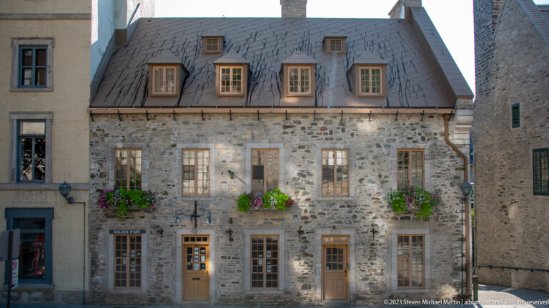 Broad pale stone house with steep gray roof, four dormers, tall multi-pane windows, three doors, and bright flower boxes.