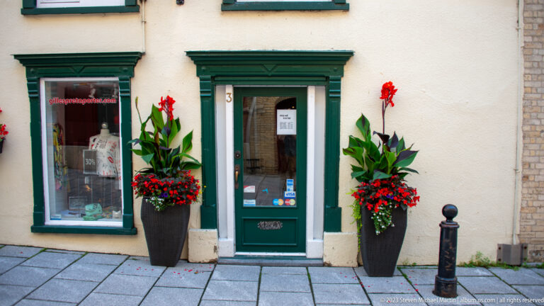 Small pale storefront with dark green door and window trim, door number 3, and tall planters filled with red flowers.