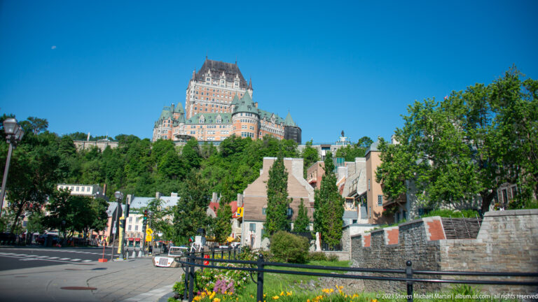 Château Frontenac on the bluff above a sloping street lined with smaller buildings, stone walls, gardens, and trees.