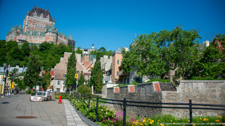 Descending street bordered by flowers, railing, trees, and low buildings, with Château Frontenac visible high above the bluff.
