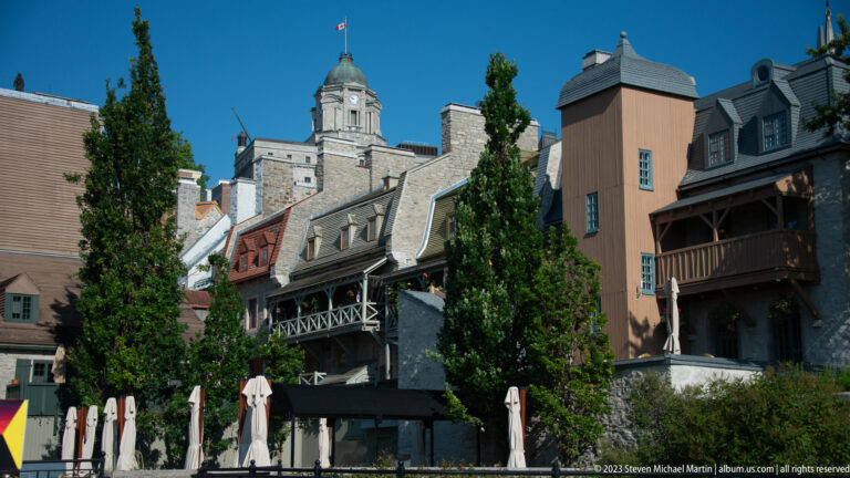 Clustered historic buildings with steep roofs, wooden galleries, chimneys, café umbrellas, and a domed flag-topped tower behind.