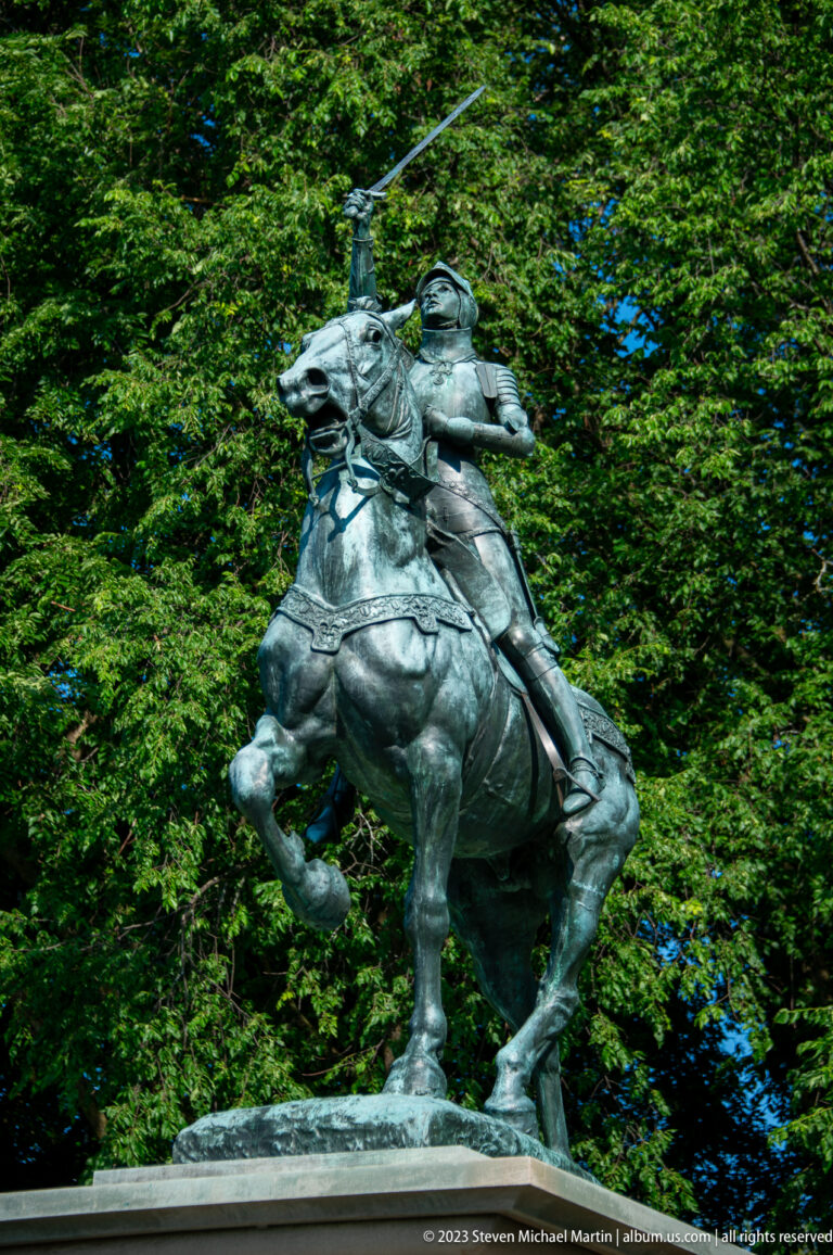 Bronze armored rider raising a long sword while mounted on a rearing horse against dense green trees.