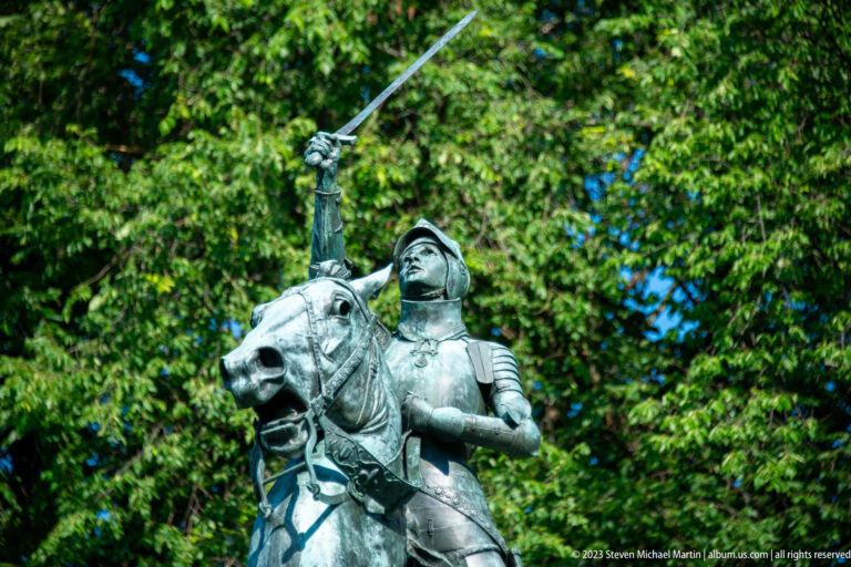 Close upward view of a bronze armored rider on horseback, raising a long sword above the horse’s bridled head.