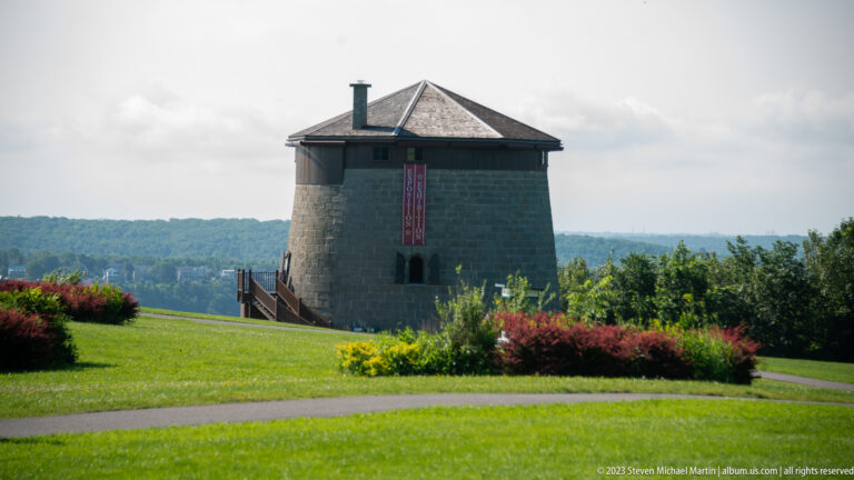 Round stone defensive tower with broad roof, narrow openings, and raised entrance platform amid lawns and flower beds.