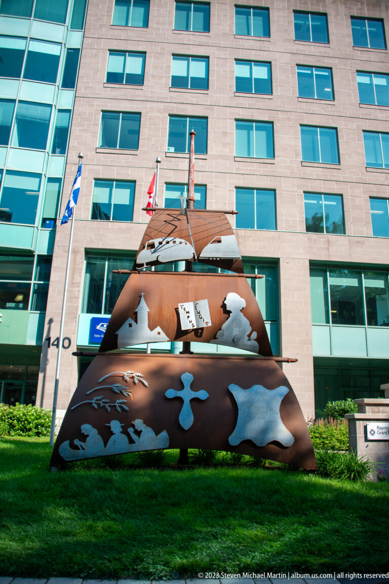 Large rust-colored pyramidal steel sculpture with curved cutouts and pale inset forms on a lawn before a modern building.