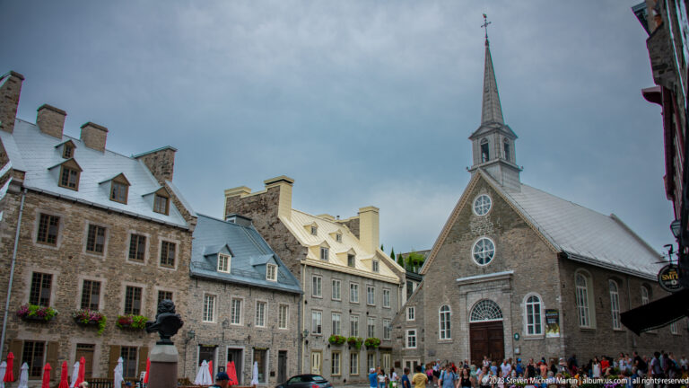 Notre-Dame-des-Victoires at Place Royale, a small stone church with steep roof and spire, framed by historic buildings and visitors.