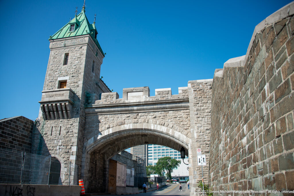 Massive stone arched gateway beneath a square tower and crenellated wall, with a modern road passing through the fortifications.