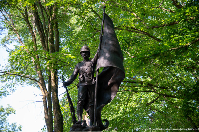 Bronze soldier in period military dress standing beneath trees while holding a large flag and long weapon.