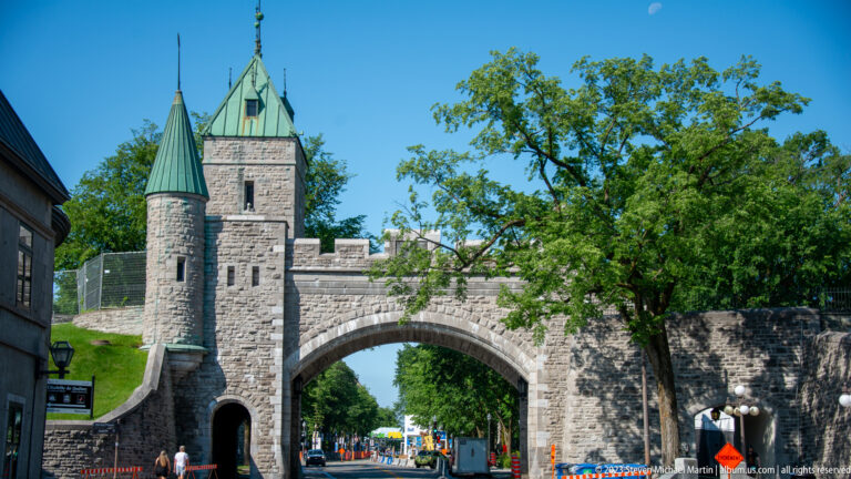 Stone fortified gateway with round turret, square tower, green roofs, crenellated walls, roadway, and grassy ramparts.